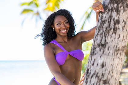 Aniya Harvey wearing a purple bikini standing next to a tree.