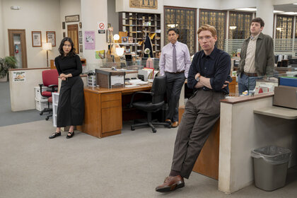 Nicole (Ramona Young), Oscar Martinez (Oscar Nuñez), Ned (Domhnall Gleeson), and Adam (Alex Edelman) standing in by their desks in The Paper Season 2 Episode 7.