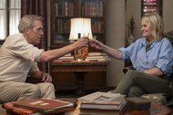 Neville (Hugh Laurie) and Elise (Amy Poehler) sit across from each other in a living room, raising glasses in a toast beside a table with books and a lamp.