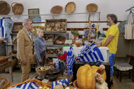 Neville (Hugh Laurie), and Elise (Amy Poehler) stand facing Lambros (Jason Mantzoukas) inside a crowded souvenir shop filled with baskets and Greek flags.