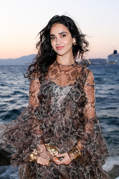 Geraldine Viswanathan poses by the water at sunset wearing a patterned sheer dress and holding a metallic clutch.