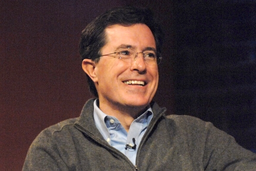 Stephen Colbert wearing a gray sweater sitting on a panel