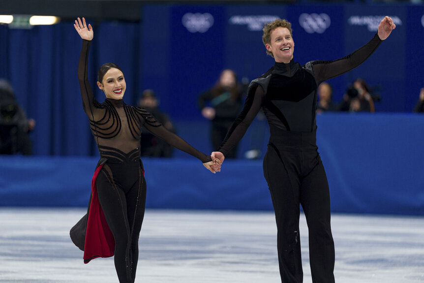 Madison Chock and Evan Bates wave while on the ice at the 2026 Milan Cortina Olympics.