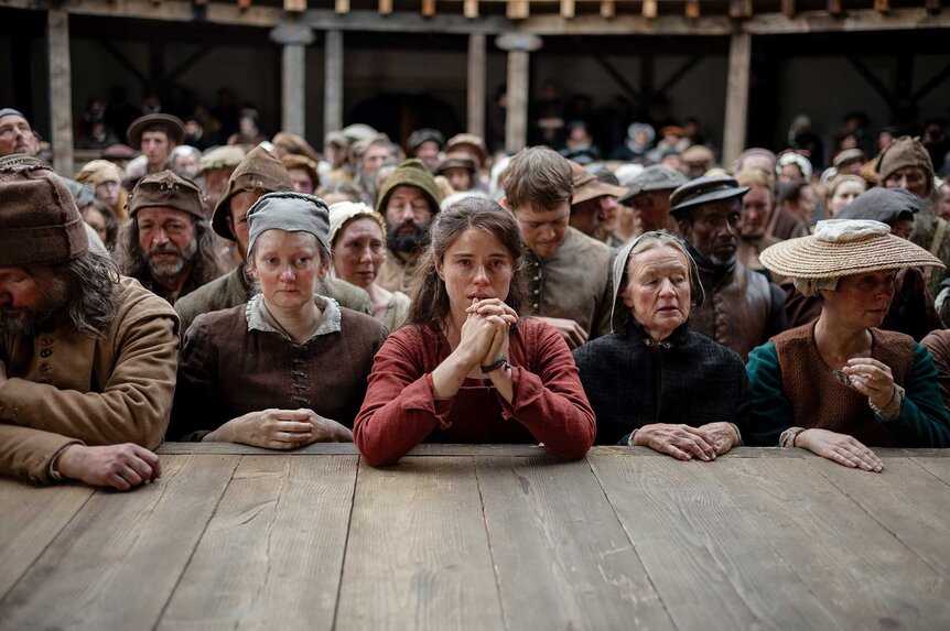 Agnes leaning against a stage with Bartholomew and other audience members in Hamnet (2025).