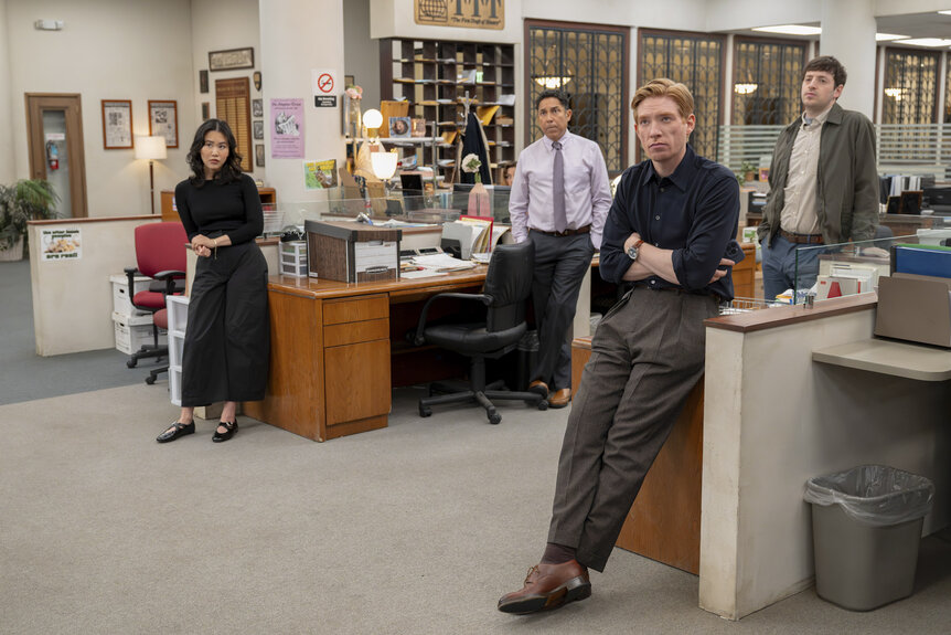 Nicole (Ramona Young), Oscar Martinez (Oscar Nuñez), Ned (Domhnall Gleeson), and Adam (Alex Edelman) standing in by their desks in The Paper Season 2 Episode 7.