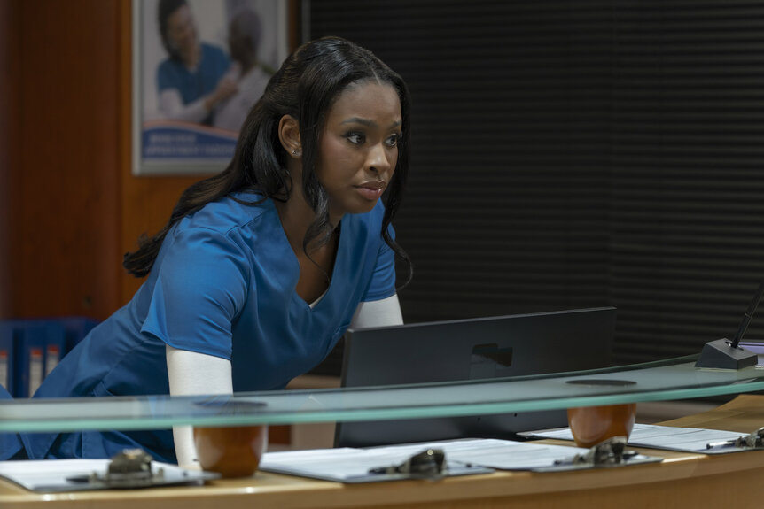Jasmine (Coco Jones) standing behind a reception desk in Strung (2026).