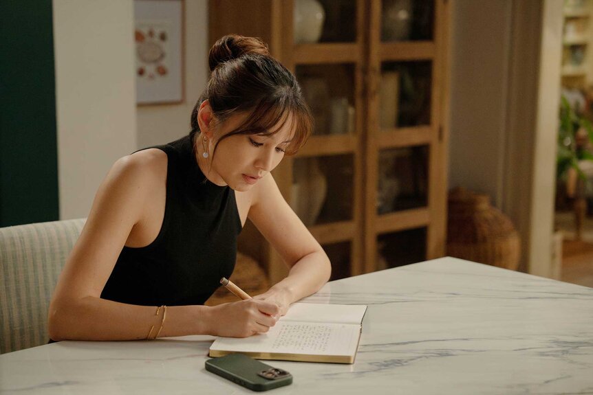 Gigi sitting at a table and writing in The Five Star Weekend Season 1.