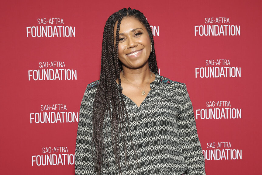 Toks Olagundoye posing in front of a step and repeat.